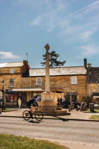 A cyclist rides past a historic monument in a picturesque English village on a sunny day.
