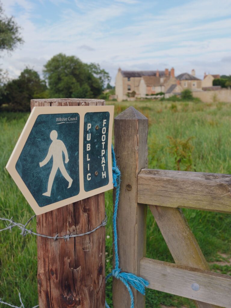 A public footpath sign along a rustic path with a traditional English countryside backdrop.